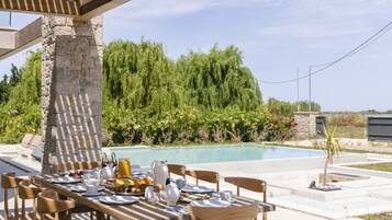 An outdoor dining area with a wooden pergola, a stone pillar, and a pool in the background.
