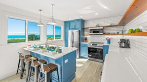 Een moderne keuken met een groot eiland, blauwe kastjes, roestvrijstalen apparatuur en uitzicht op zee.