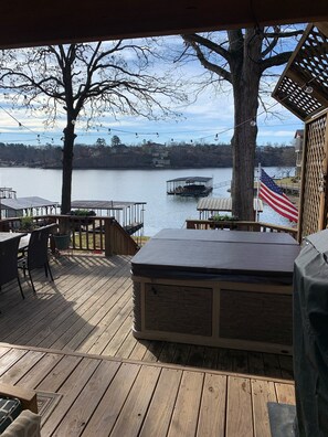 A wooden deck with a hot tub, outdoor seating, and a view of a lake with a dock and an American flag. - Cottage -Private Hot Tub, Fire Pit, Covered Deck, 20 steps from Lake Hamilton (Hot Springs)
