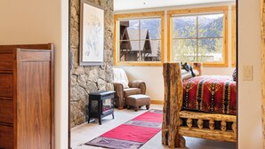 Quarto com parede de pedra, cama de madeira, uma cadeira, uma mesinha e janela com vista para as montanhas.