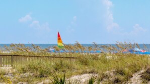A beach with a flag, beach umbrellas, and grass in the foreground. - Brigantine Quarters, Shipyard Plantation, Hilton Head 2 bed/2 bath condo (Hilton Head)