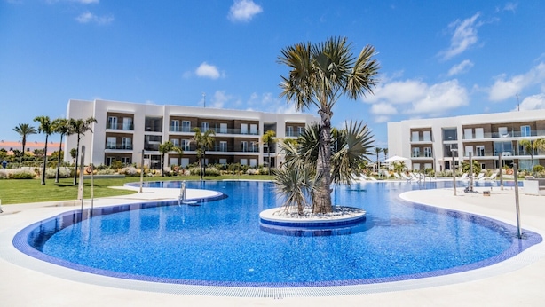Une piscine d’un complexe hôtelier, bordée de palmiers, avec un ciel bleu azur et des bâtiments modernes en arrière-plan.