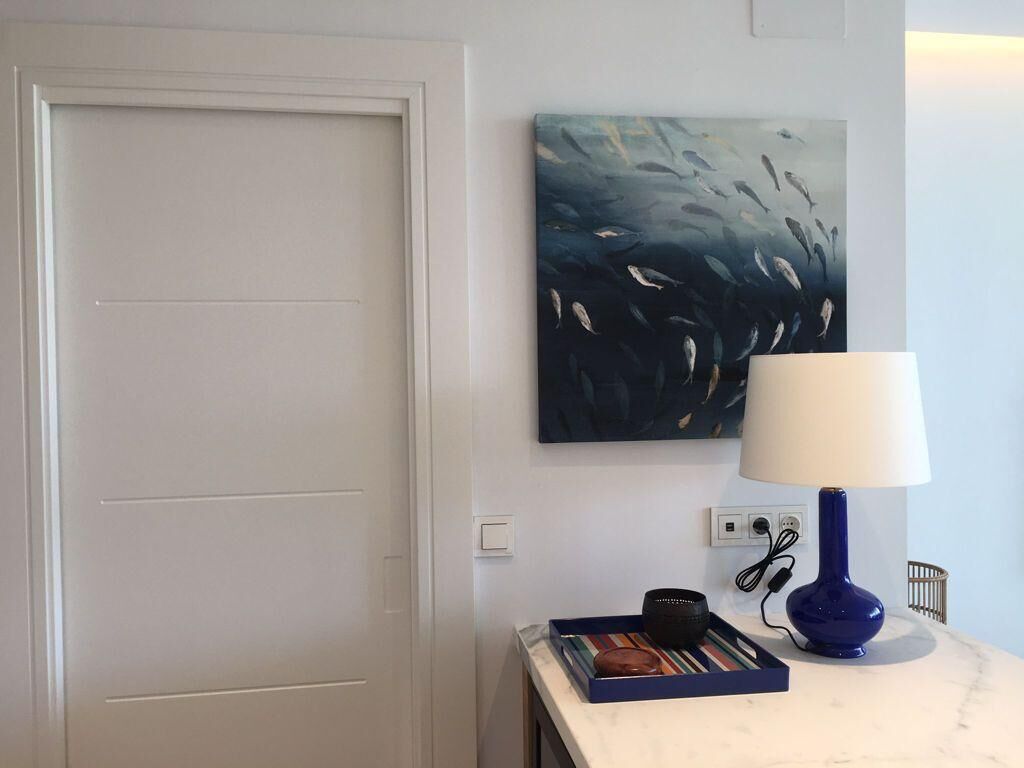 A room with a white door, a wall-mounted painting of fish, a blue lamp, and a small table with a book and a black object.