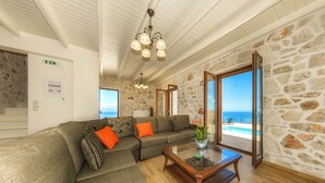 A spacious living area with a large sectional sofa, a glass coffee table, and a view of the ocean through large windows. - Orient Villas Deluxe (Zakynthos)