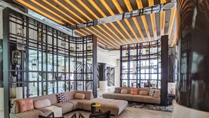 Uma sala de estar ampla com um grande sofá modular, duas poltronas e uma mesa de centro. O espaço apresenta um design moderno com teto de madeira e grandes janelas que permitem entrada de luz natural.