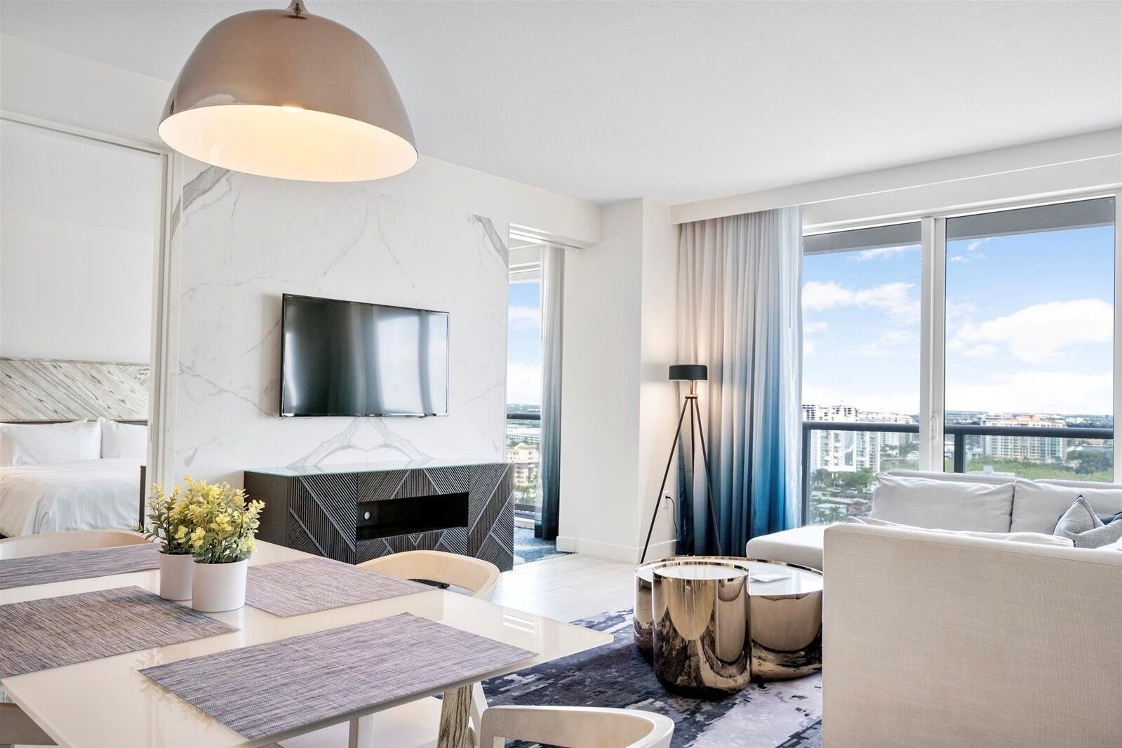 Habitación de hotel moderna con un ventanal grande, televisor de pantalla plana, zona de comedor con mesa y sillas, y un sofá.