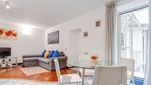 A modern living room with a glass dining table, white chairs, a grey sofa with blue and yellow cushions, a flat-screen TV, and artwork on the walls. - Casa Querida Central and Elegant Flat (Naples)