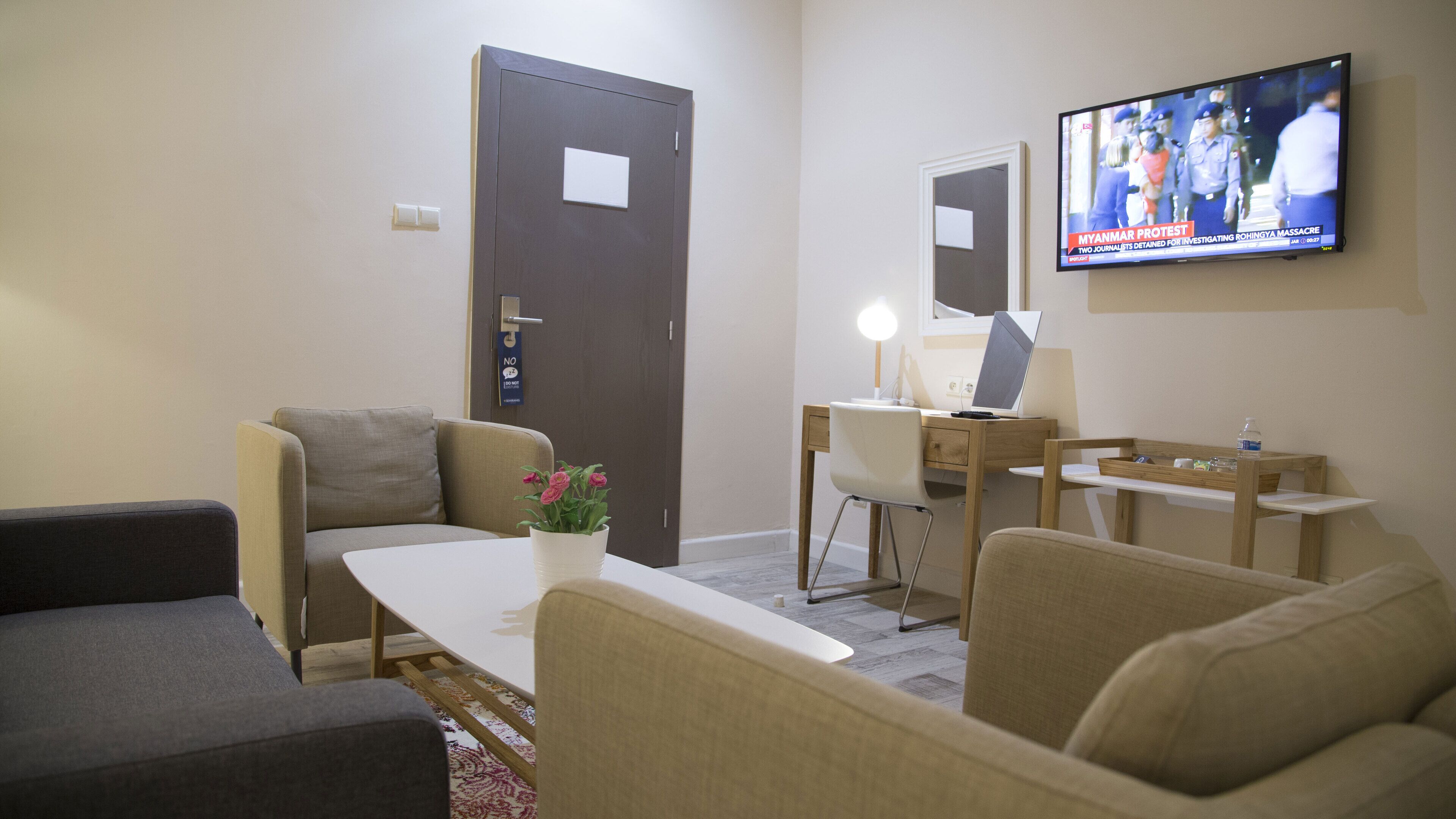Ein Hotelzimmer mit einer Couch, einem kleinen Tisch mit Blumen in einer Vase, einem Schreibtisch mit einem Laptop und einem an der Wand montierten Fernseher.