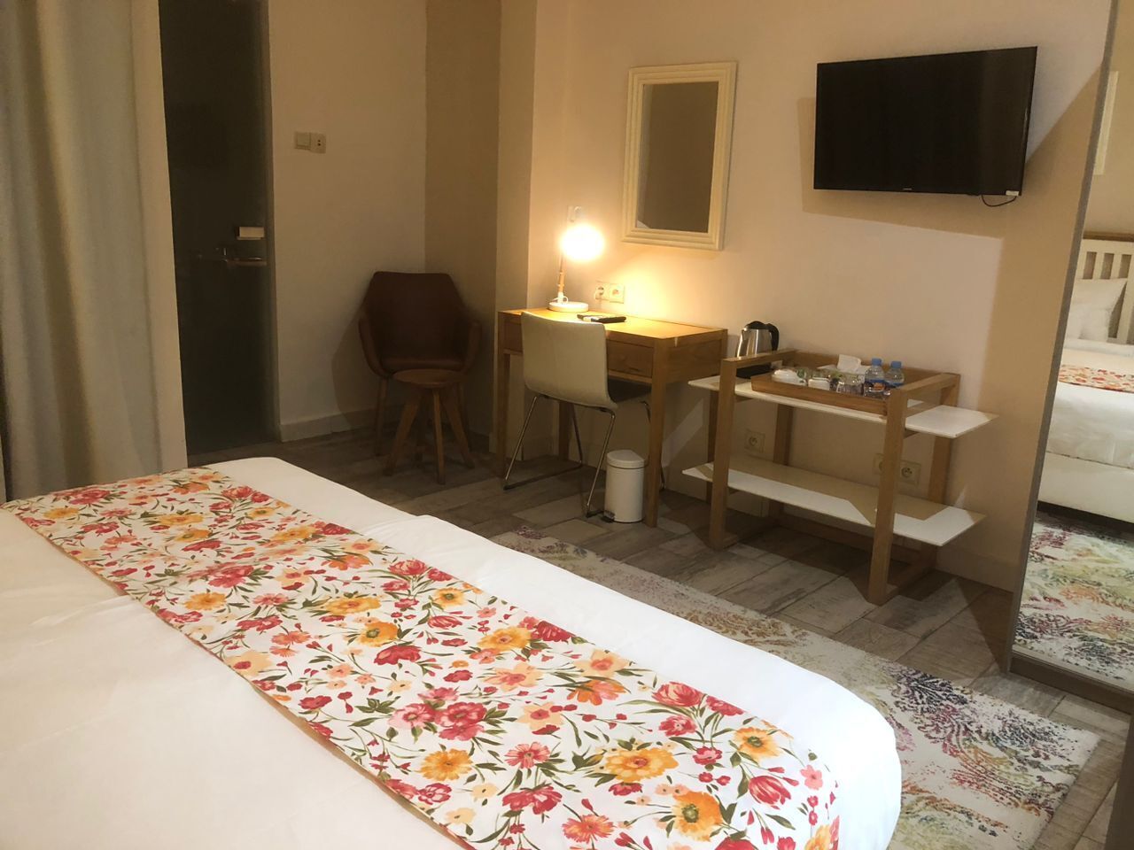 A hotel room with a bed, a desk with a lamp, a chair, a TV, and a small table with items on it.