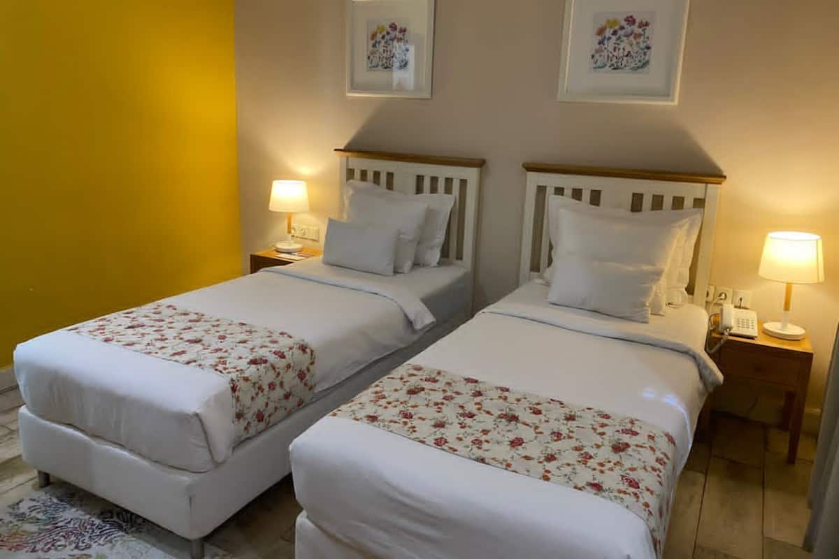 Duas camas de solteiro com roupa de cama branca e estampas florais, mesas de cabeceira com abajures, e quadros com motivos florais nas paredes.