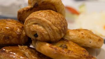 A plate of pastries, including croissants and puff pastries, on a table with a glass of orange juice and a sandwich in the background.