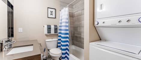A modern bathroom with a built-in washing machine, a shower with a blue and white striped curtain, a toilet, and a sink with a wooden countertop.