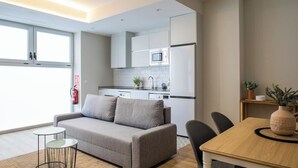 A modern apartment with a grey sofa, a small round table, a wooden dining table, and a kitchen area with white cabinets and appliances. - Port Suite X | 4 Guests  | AC & WiFi | 15 min Ciudad Artes (Valencia)