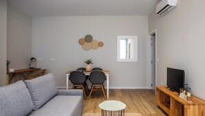 A modern living room with a grey sofa, a dining table with chairs, a small round table, a TV on a wooden stand, and an air conditioning unit on the wall. - Port Suite X | 4 Guests  | AC & WiFi | 15 min Ciudad Artes (Valencia)