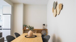 A modern kitchen with a white refrigerator, a wooden dining table with four chairs, a potted plant, and a small side table with a lamp. - Port Suite X | 4 Guests  | AC & WiFi | 15 min Ciudad Artes (Valencia)