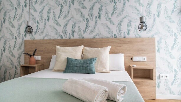 A neatly made bed with a wooden headboard, two bedside tables, and a wall with a leafy pattern. - Port Suite X | 4 Guests  | AC & WiFi | 15 min Ciudad Artes (Valencia)