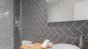 A modern bathroom with a herringbone tile pattern, a wooden vanity, a white sink, and a glass shower enclosure. - Port Suite V | 4 Guests | AC & WiFi | 15 min Ciudad Artes (Valencia)