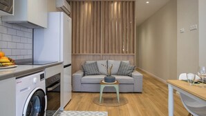 Ein kompakter Wohnbereich mit Küchenzeile, einem grauen Sofa mit Kissen, einem kleinen Holztisch und einem Essbereich mit Tisch und Stühlen.