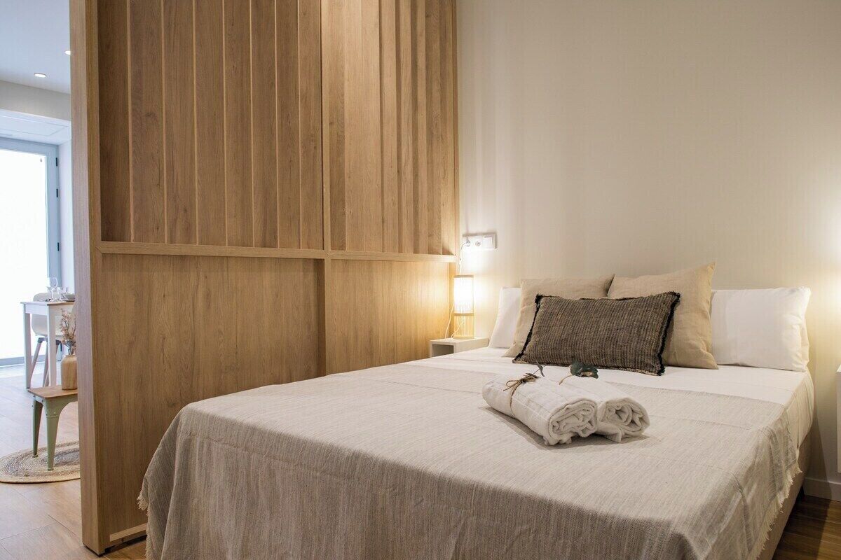 Een keurig opgemaakt bed met een witte spreideken en twee kussens, een wand van houten panelen en een klein nachtkastje met een lamp.