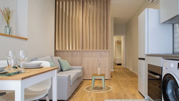 A modern kitchen with a washing machine, a dining area with a table set for two, and a living area with a sofa and a decorative wall panel. - Port Suite II | 4 Guests | AC & WiFi | 15 min Ciudad Artes (Valencia)