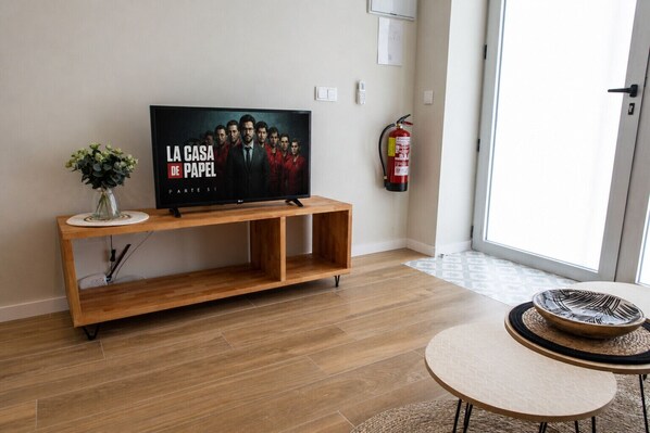A living room with a flat-screen TV on a wooden stand, a small table with a woven table runner, and a fire extinguisher mounted on the wall.