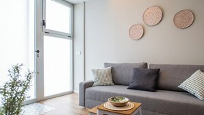 Een moderne woonkamer met een grijze bank, een klein houten tafeltje met een schaal, en een groot raam met uitzicht op groen.