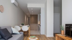 Een moderne woonkamer met een grijze bank, een houten salontafel en een televisie op een houten dressoir. Op de achtergrond is een eethoek met een tafel en stoelen zichtbaar.