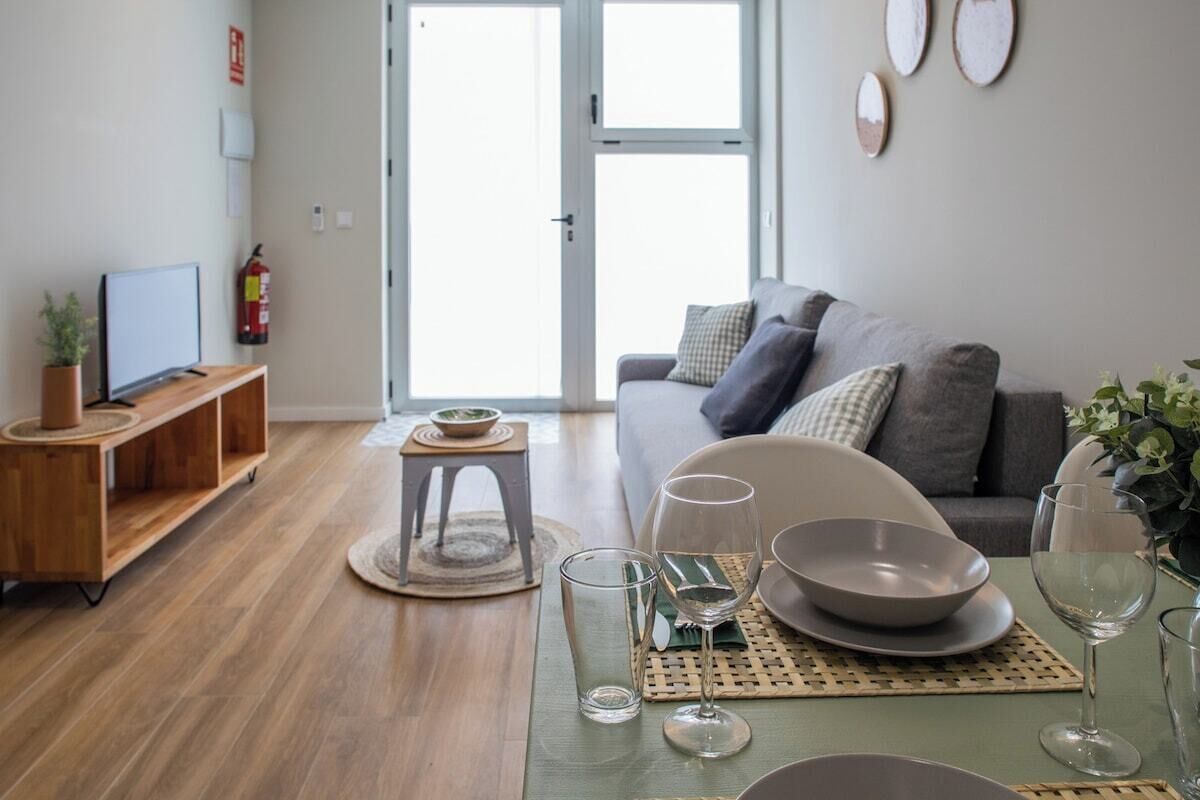 Een moderne woonkamer met een grijze bank, een houten tv-meubel en een eettafel gedekt met kommen en wijnglazen.