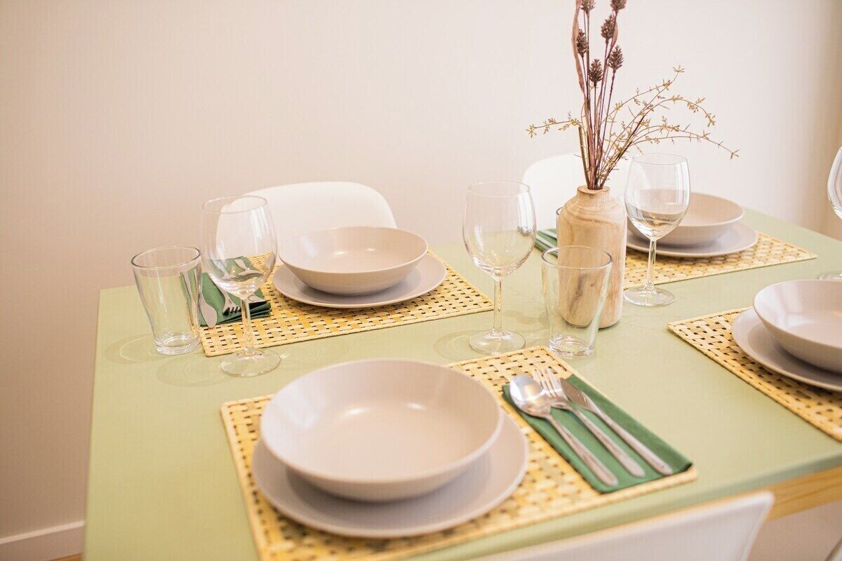 Une table à manger dressée avec de l’assiette, des bols et des verres blancs, agrémentée d’un vase de fleurs séchées et d’un set de table vert.