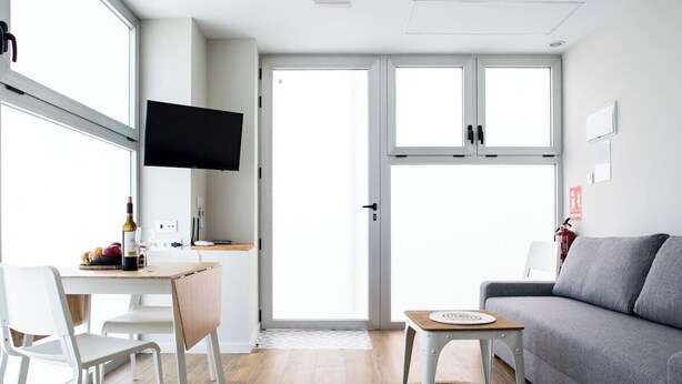 Una sala de estar moderna con un sofá gris, una mesa de comedor de madera con sillas, un televisor de pantalla plana montado en la pared y un amplio ventanal corredizo que da a un espacio exterior.
