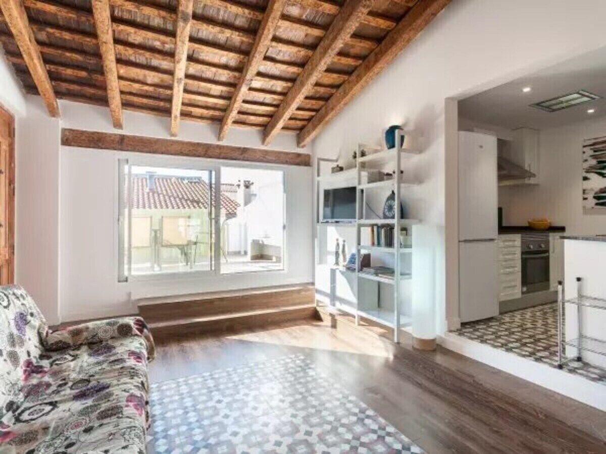 A spacious living area with a wooden ceiling, a patterned rug, a sofa, and a kitchen with a tiled backsplash.