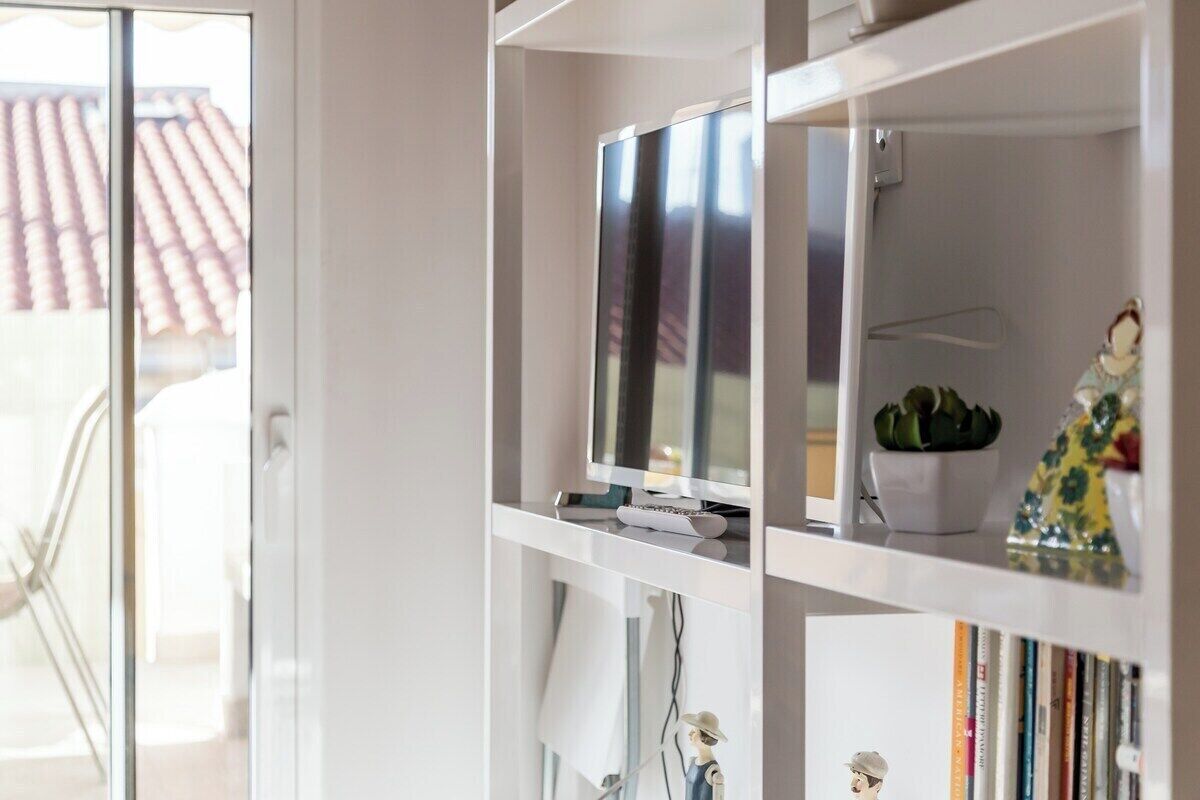 Een moderne woonkamer met een glazen deur naar een balkon, een flatscreen televisie op een witte plank, en een kleine plant in een pot.