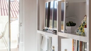 Una sala de estar moderna con un ventanal que da a un balcón, un televisor de pantalla plana sobre una estantería blanca y una pequeña planta en maceta.