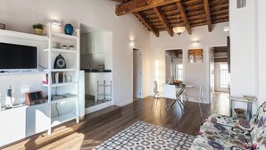 Una sala de estar moderna con techo de madera, estanterías blancas, alfombra estampada y un comedor con sillas y una mesa.