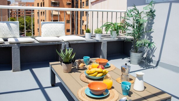 Un balcón con una mesa dispuesta para el desayuno, que incluye frutas, una cafetera y vistas de la ciudad.