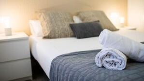 Una cama bien hecha con una toalla enrollada al lado, una lámpara de noche y una mesita de noche con cajón.