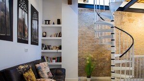 Una sala de estar moderna con un sofá de cuero negro, estanterías blancas con libros, una planta en maceta y una escalera en espiral frente a una pared de ladrillo.