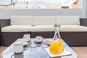 Un coin petit-déjeuner aménagé sur un balcon, avec un canapé en osier, des tasses et des verres blancs, et une carafe de jus d’orange.