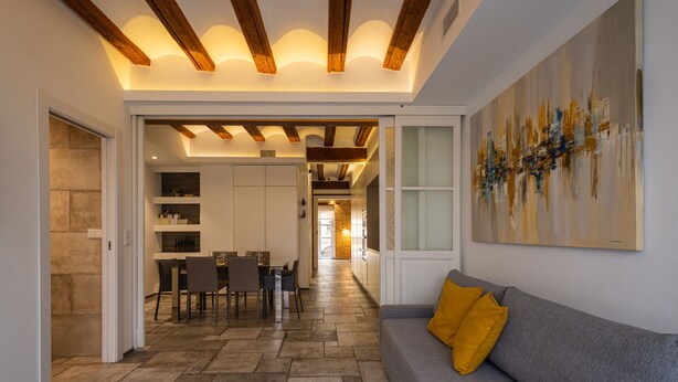 Een modern interieur met een stenen vloer, een grijze bank met een geel kussen, een eethoek met stoelen en een tafel, en een keuken op de achtergrond.