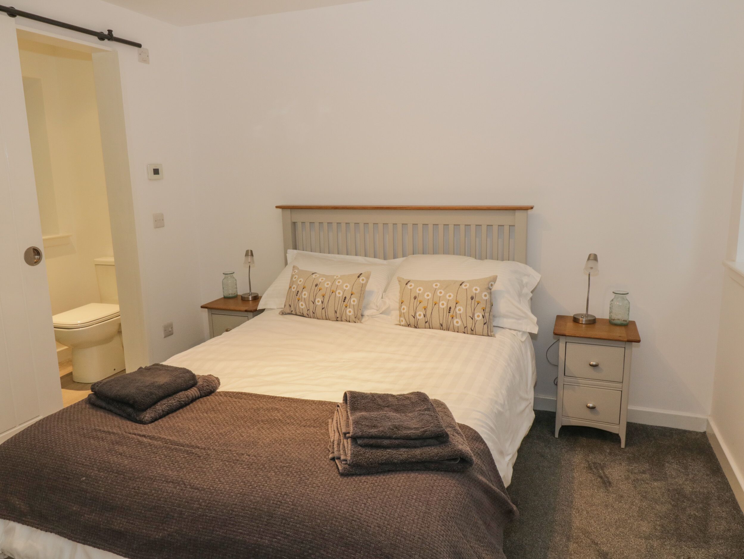 A bedroom with a bed, two bedside tables, a nightstand, a wooden headboard, and a door leading to a bathroom.