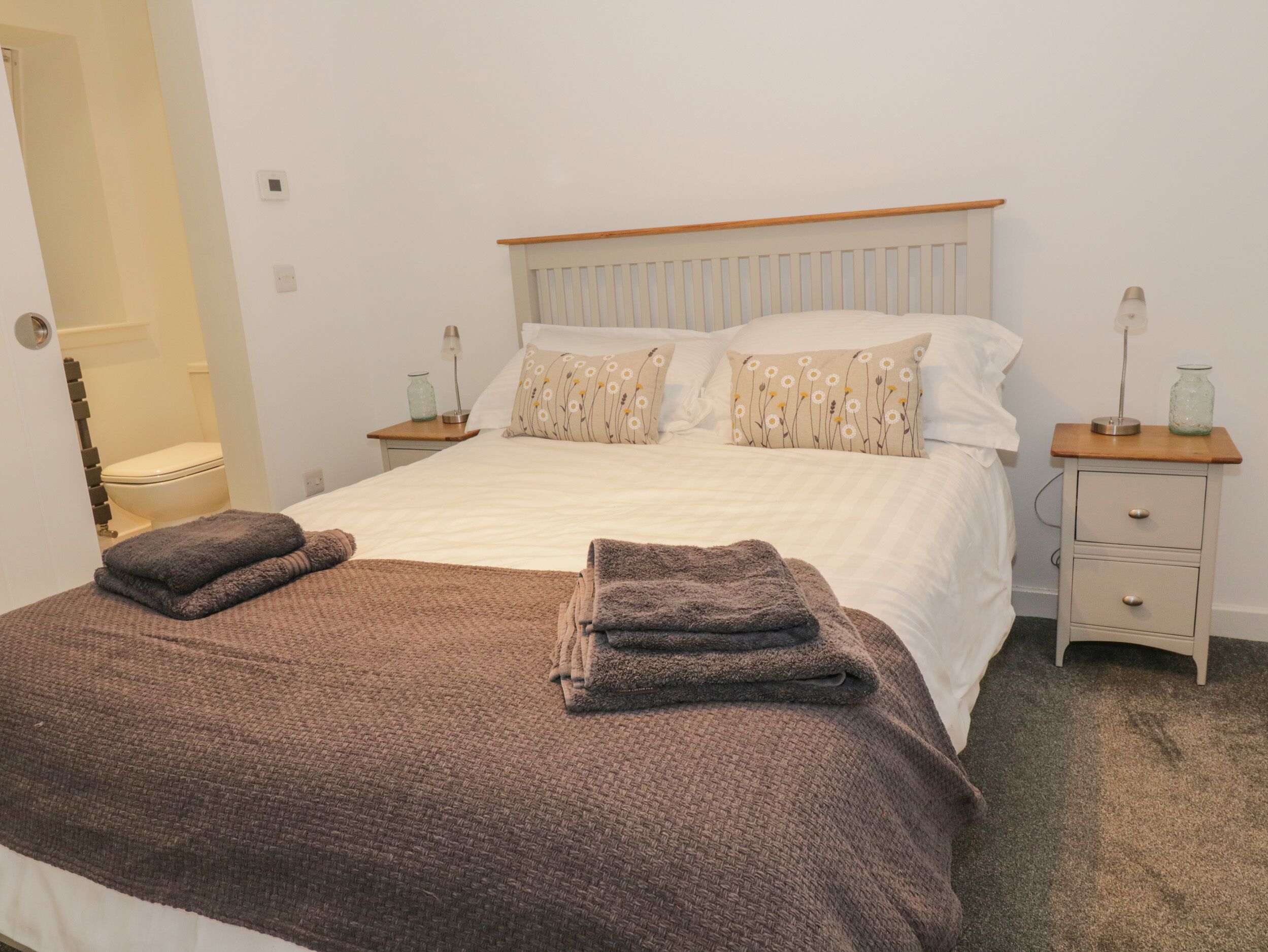 A neatly made bed with a brown blanket and two folded towels, a bedside table with a lamp, and a small drawer unit.