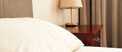 A neatly made bed with white linens, a wooden nightstand with a lamp, and a curtain in the background.