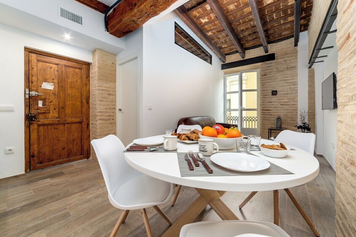 Un comedor con una mesa redonda, sillas blancas y una disposición para el desayuno que incluye frutas y pasteles.