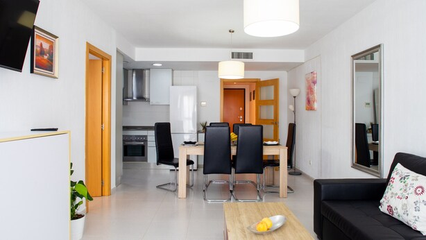 Ein modernes Wohnzimmer mit einem schwarzen Sofa, einem Couchtisch mit Früchten und einem Essbereich mit Holztisch und schwarzen Stühlen.