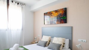 Ein ordentlich bezogenes Bett mit einer grünen Tagesdecke und mehreren Kissen, ein Bild an der Wand und ein Fenster mit transparenten Vorhängen.