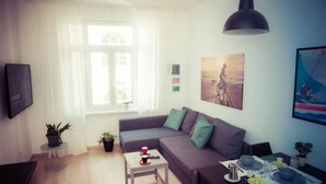 Un salon moderne comprenant un canapé, une table à manger pour deux personnes, une table basse et une grande fenêtre ornée de rideaux légers.