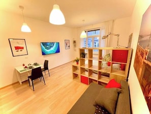 A modern living room with a wooden floor, a grey sofa, a dining table with chairs, a TV, and a bookshelf.