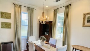 Un espace repas comprenant une table en bois, des chaises blanches, un lustre et une vue sur l’extérieur grâce à de grandes fenêtres.