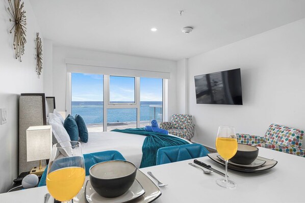 A modern hotel room with a large bed, a dining table with breakfast items, a TV, and a view of the sea.