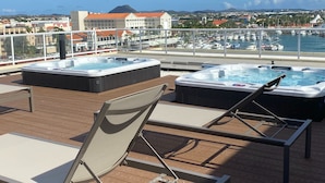 Une terrasse dotée de deux bains à remous et de chaises longues, offrant une vue sur une marina et une ville côtière.